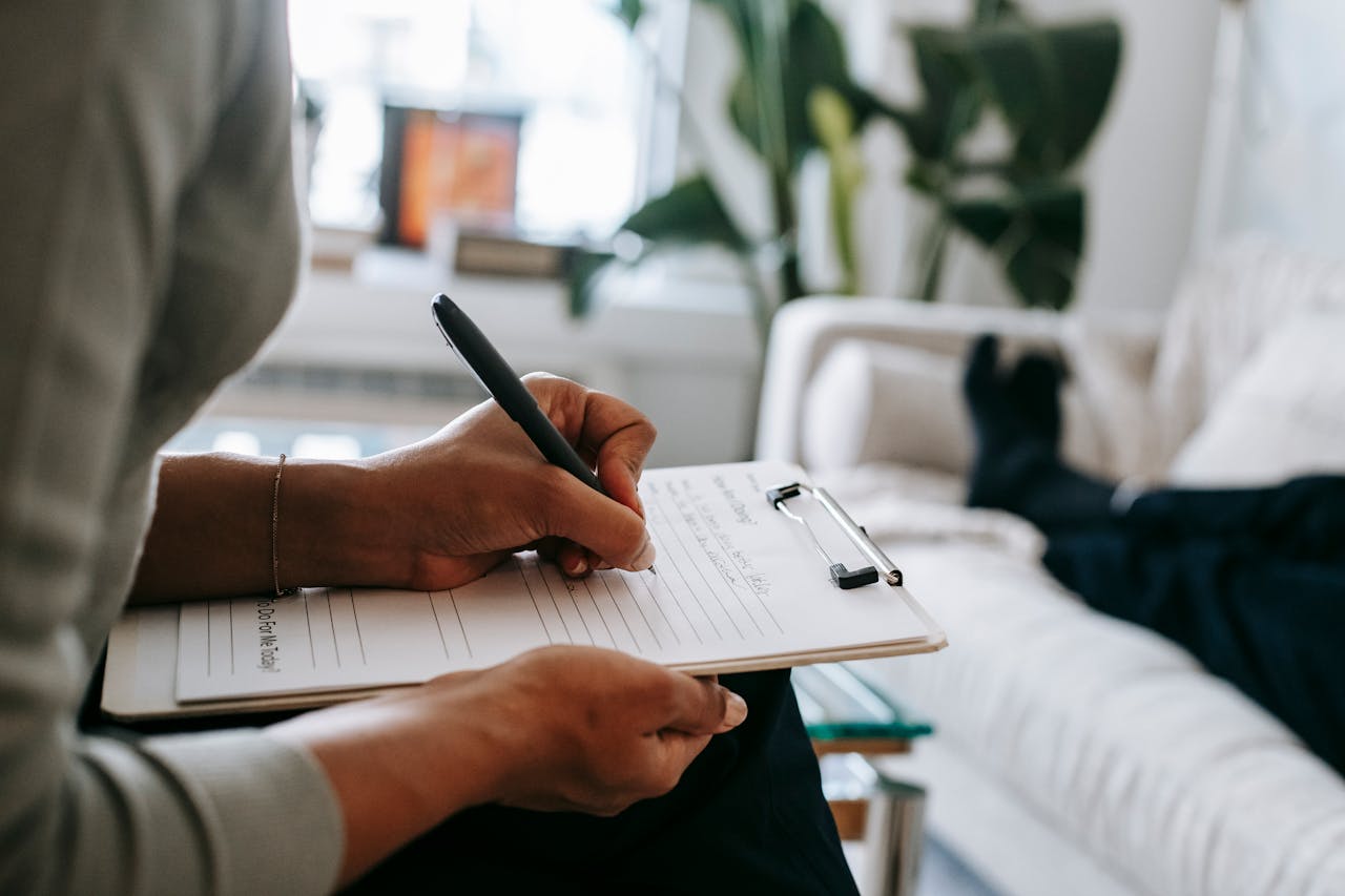 about-03 Unrecognizable ethnic female therapist taking notes on clipboard while filling out form during psychological appointment with anonymous client lying on blurred background