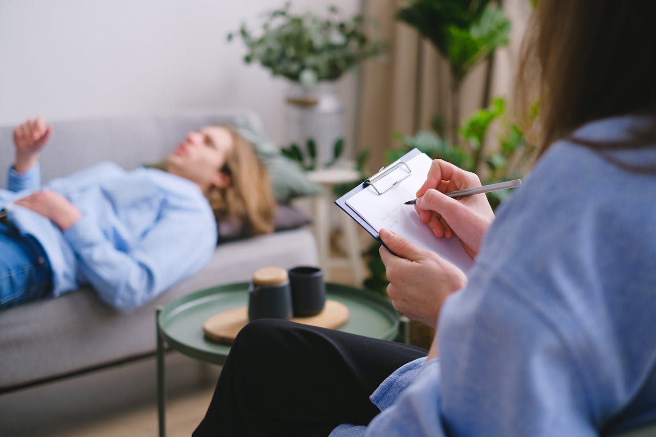services-02 Unrecognizable professional therapist writing on clipboard while sitting against male client lying on couch on blurred background during mental health therapy